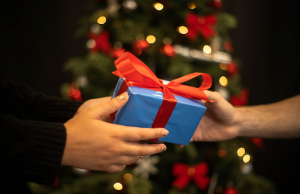 Person handing a gift to someone in front of a Christmas tree
